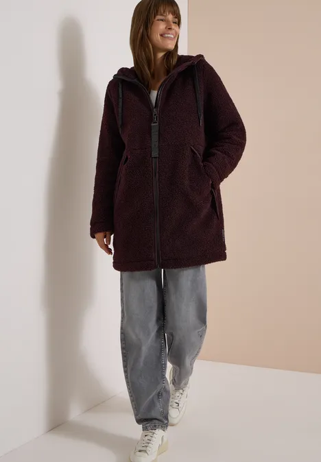 Strukturmix Teddy-Mantel mit Kapuze berry red Frau trägt burgunderfarbenen Teddy-Mantel, graue Jeans und weiße Sneaker, lächelnd vor hellem Hintergrund.