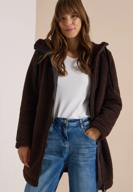 Frau in braunem Teddy-Mantel mit Kapuze über weißem T-Shirt und Jeans, lächelt leicht.