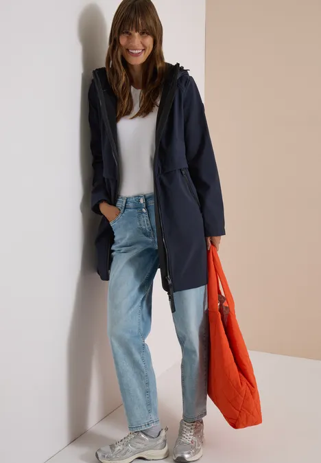 Frau lehnt an Wand, trägt dunkelblauen Mantel, weißes Shirt, helle Jeans, weiße Sneaker und eine orangefarbene Tasche