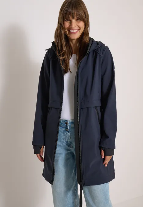 Frau in langer dunkelblauer Kapuzenjacke, weißem Shirt und Jeans lächelnd.