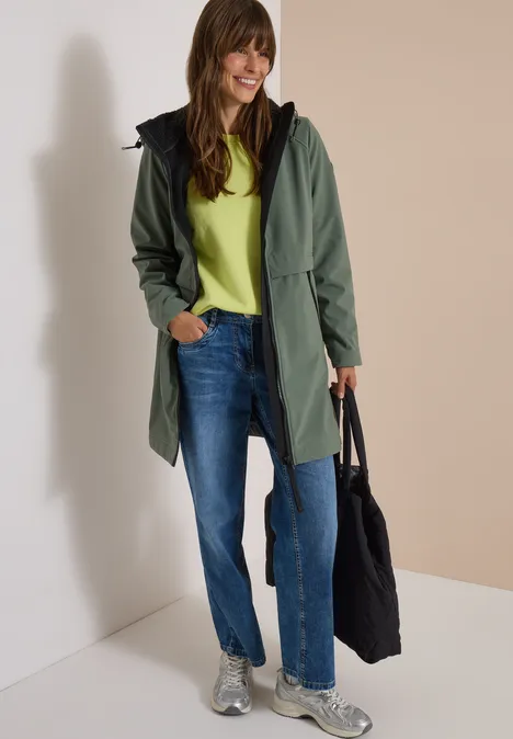 Eine lächelnde Frau in grünem Mantel, Jeans und Sneakern hält eine schwarze Tasche.