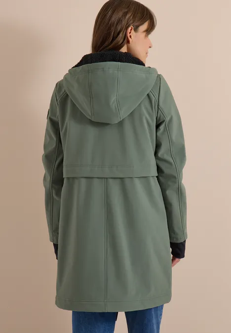 Eine Person steht mit dem Rücken zur Kamera und trägt einen grünen, knielangen Parka mit Kapuze.