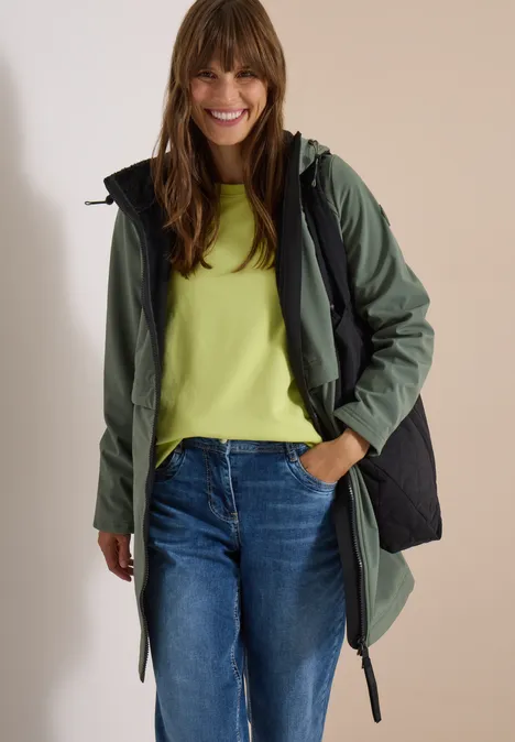 Eine lächelnde Frau in Jeans, grünem Shirt und dunkelgrünem Parka vor hellem Hintergrund.