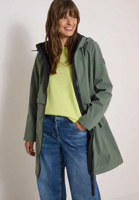 Frau in grünem Regenmantel mit Kapuze, gelbem Shirt und blauen Jeans vor weißem Hintergrund.
