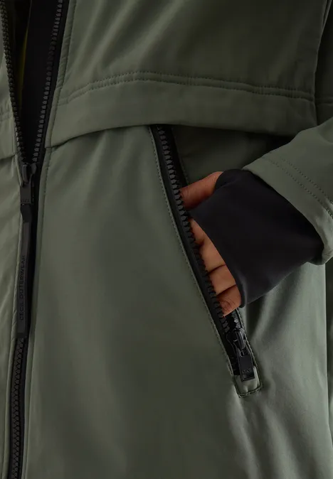 Hände stecken in der Reißverschlusstasche einer olivgrünen Jacke mit schwarzen Bündchen.