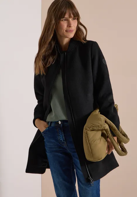 Vrouw draagt zwarte jas, jeans en groen shirt, houdt een kaki jas in haar hand