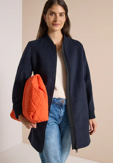 Modische Frau trägt marineblauen Mantel, weißes Top, Jeans und hält eine orangefarbene Tasche unter dem Arm.