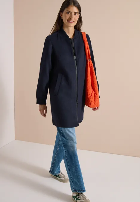 Eine Frau in einem dunklen Mantel, blauen Jeans und roter Tasche steht vor hellem Hintergrund.