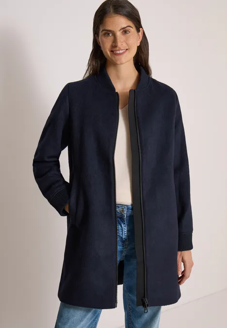 Frau in dunkelblauem Mantel mit Reißverschluss und Taschen, dazu Jeans und weißer Pullover.