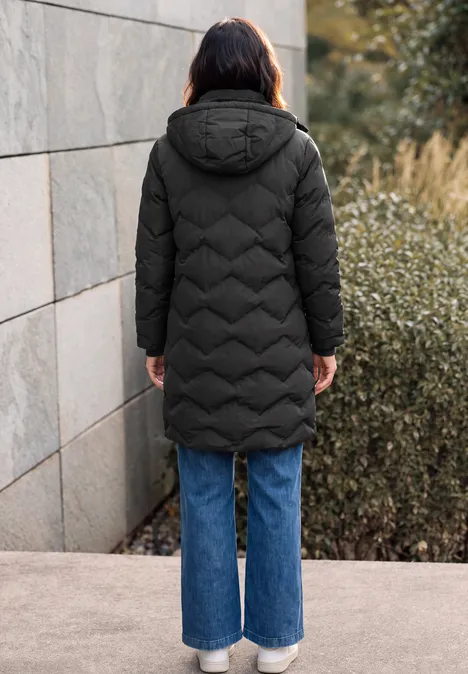 Vue de dos: personne en longue doudoune noire à capuche et jeans larges bleus devant un mur et des arbustes.