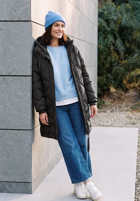 Une femme souriante en manteau d'hiver noir, bonnet bleu et jeans appuyée contre un mur extérieur.