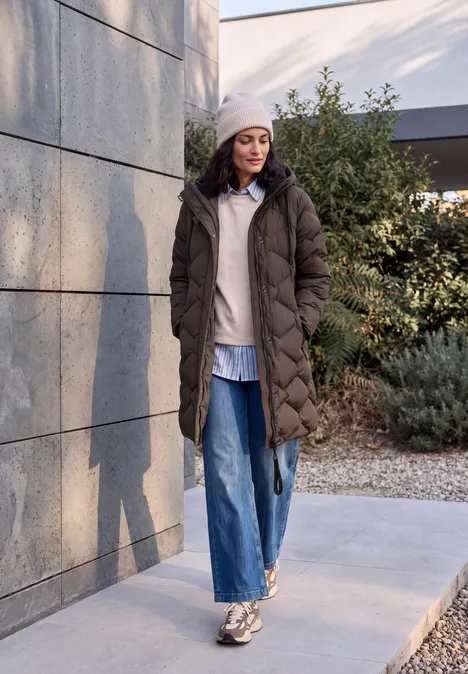 Une femme en long manteau matelassé, bonnet et jean marche à l'extérieur près d'un mur.