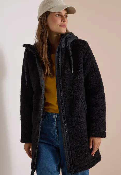 Vrouw in zwarte teddyjas met capuchon, jeans, mosterdgeel truitje en pet.