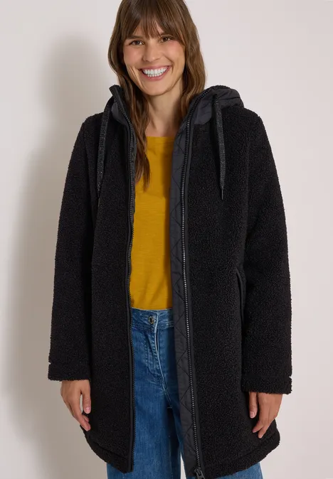 Vrouw lacht in een zwarte capuchonjas, geel shirt en spijkerbroek.