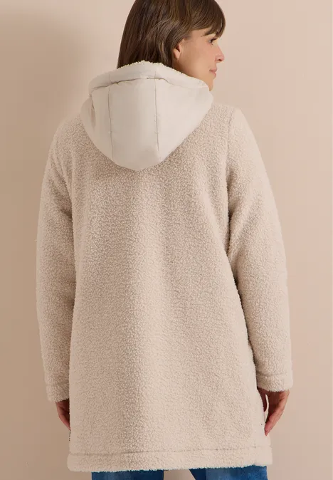 Persoon van achteren draagt een crèmekleurige teddyjas met capuchon tegen een neutrale achtergrond.