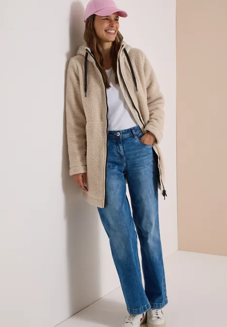 Een lachende vrouw in jas, jeans en roze pet leunt tegen een muur.