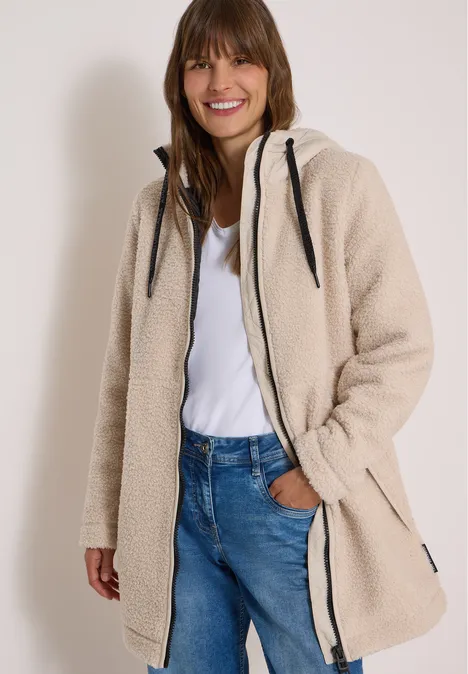 Vrouw in beige teddy-fleecejas met capuchon, jeans en wit shirt, lacht naar de camera.