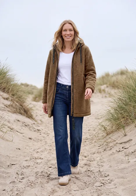 Femme en veste marron, jean et baskets, souriante, marchant sur le sable dunaire entre herbes.