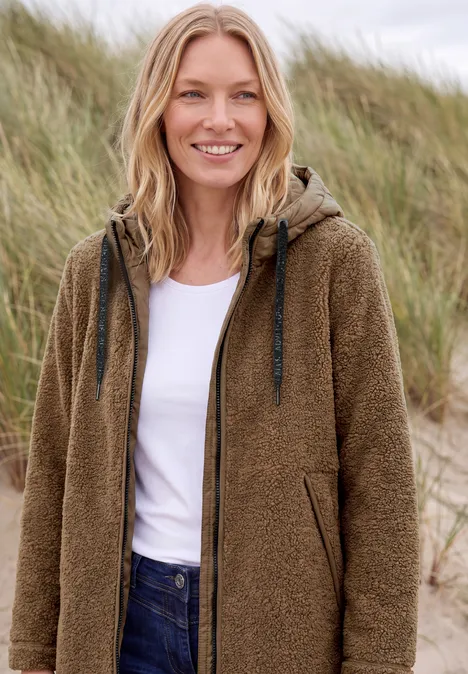 Femme souriante en veste marron en fausse fourrure et jeans, debout devant des dunes herbeuses.