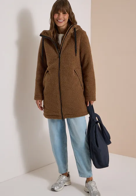 Vrouw in bruine teddyjas, baggy jeans en sneakers met donkerblauwe tas voor lichte achtergrond.