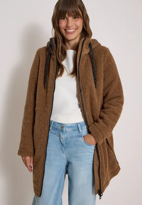 Vrouw in bruin teddyjack, wit shirt en blauwe jeans, glimlachend.