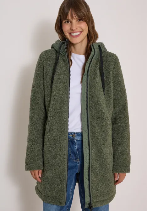 Vrouw in groene teddyjas met capuchon, wit shirt en spijkerbroek, glimlachend.