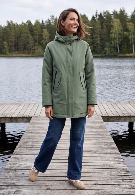 Frau in grünem Parka steht lächelnd auf einem Holzsteg am See, umgeben von Bäumen.