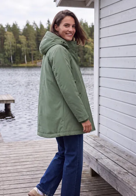 Frau in grünem Parka steht auf hölzernem Steg am See und schaut zurück.