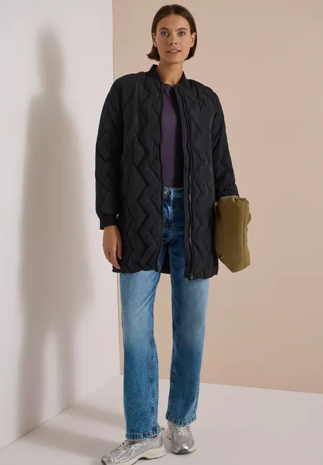 Femme en doudoune longue matelassée, tee-shirt violet, jean bleu et baskets, tenant un sac vert.