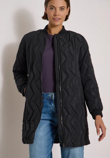 Femme en longue veste matelassée noire sur un t-shirt violet et un jean bleu, mains dans les poches.