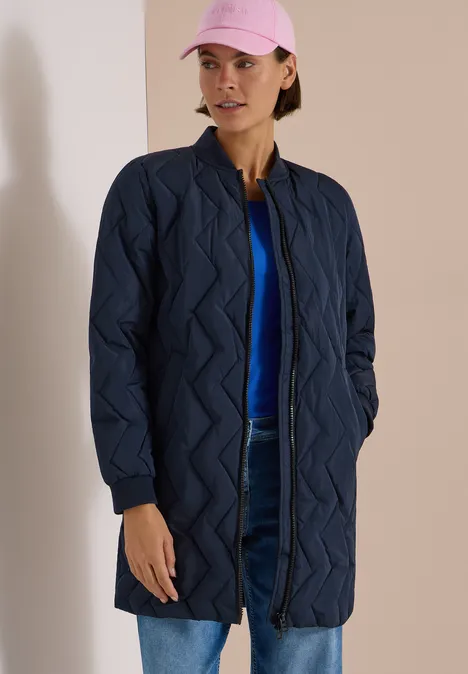 Frau trägt einen dunkelblauen Steppmantel, Jeans, blaues Shirt und rosa Cap.