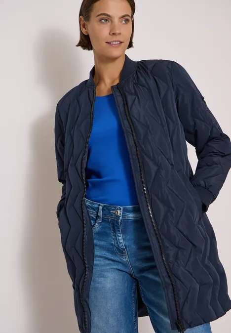 Eine Frau trägt eine lange, gesteppte dunkelblaue Jacke über einem blauen Top und Jeans.