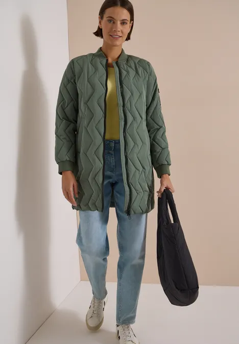 Une femme porte un manteau matelassé long vert, un jean ample, des baskets blanches et un sac noir.