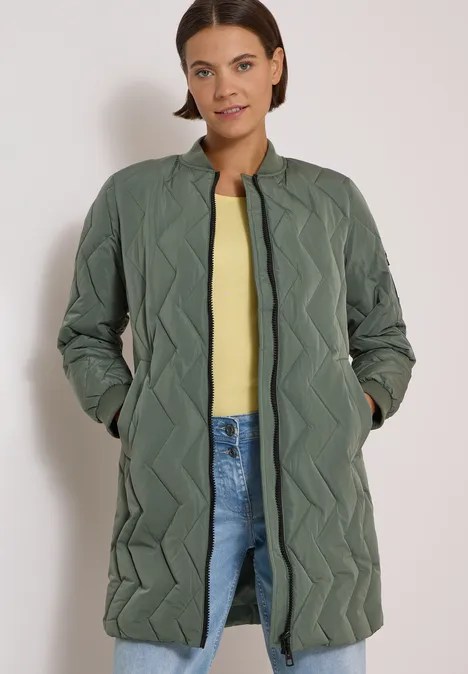 Une femme porte un manteau matelassé olive long à motif zigzag, un top jaune et un jean.