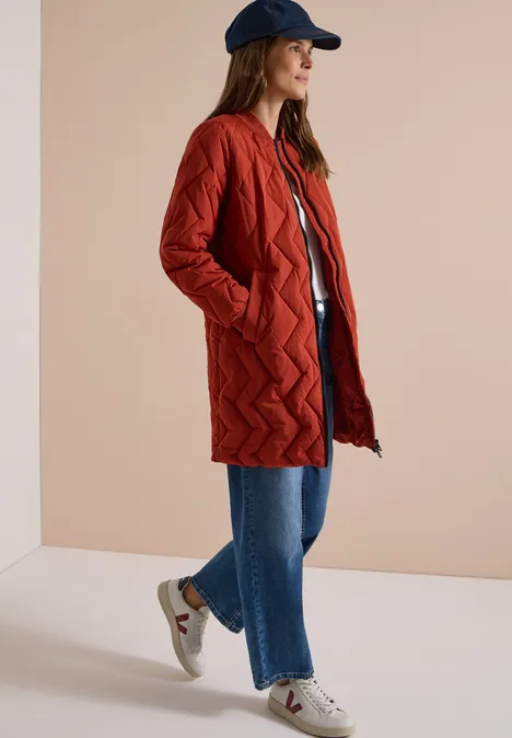Langer Blousonmantel mit Steppmuster light russet red Frau mit rotem Steppmantel, blauer Jeans, weißen Sneakern und Cap vor neutralem Hintergrund.