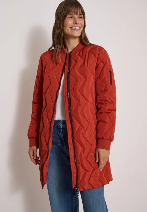 Langer Blousonmantel mit Steppmuster light russet red Frau in langem, gestepptem orangeroten Mantel mit Reißverschluss über Jeans und weißem Shirt.