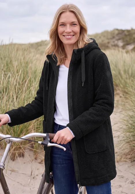 Une femme souriante en manteau sombre, debout près d’un vélo sur un paysage de dunes.