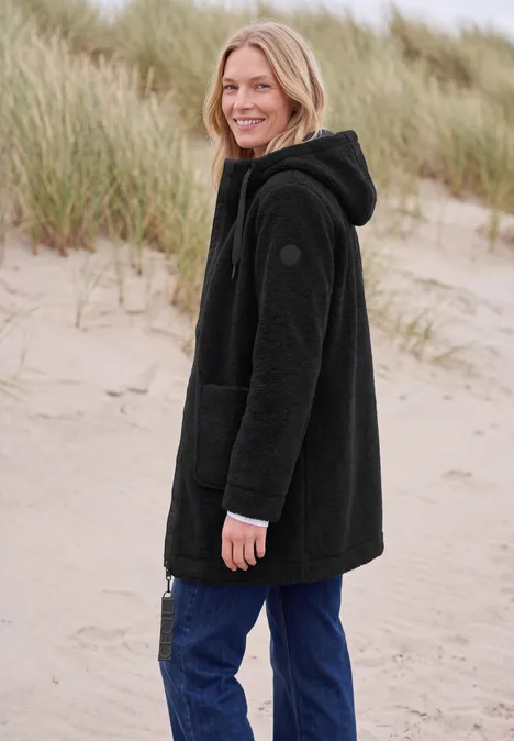Femme souriante en veste polaire noire et jean sur la plage devant des dunes.