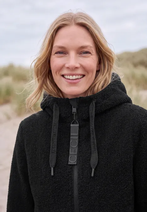 Une femme sourit en pull à capuche noir sur une plage.