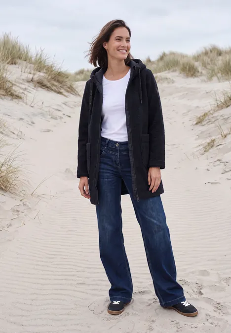 Femme en jean, t-shirt blanc et veste noire souriante, debout sur des dunes sablonneuses avec des oyats.