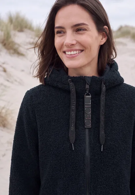 Femme souriante en sweat polaire noir devant des dunes de sable.