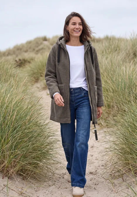 Frau in grünem Mantel und Jeans geht lächelnd durch Dünenlandschaft am Strand.