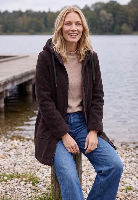 Eine Frau in brauner Jacke sitzt entspannt auf Steinen am Ufer, im Hintergrund ein Steg und ruhiger See.