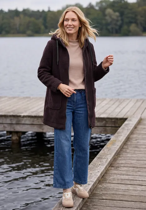Frau mit langer Jacke, Jeans und Sneakern spaziert lächelnd auf einem Steg am See.