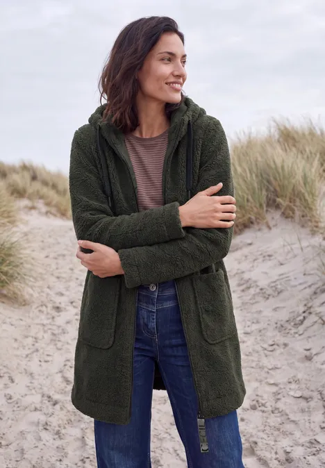 Mantel in angerauter Wolloptik olive night Frau lächelt in grüner Kapuzenjacke am Strand mit Dünen im Hintergrund.