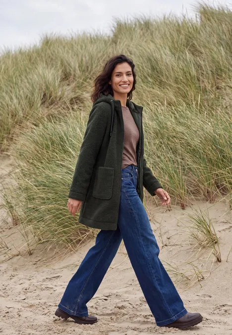 Mantel in angerauter Wolloptik olive night Eine lächelnde Frau in grünem Mantel und blauen Jeans spaziert über einen sandigen Dünenweg.