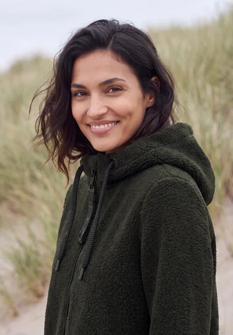 Mantel in angerauter Wolloptik olive night Eine lächelnde Frau in grünem Fleece-Hoodie vor Dünenkulisse.