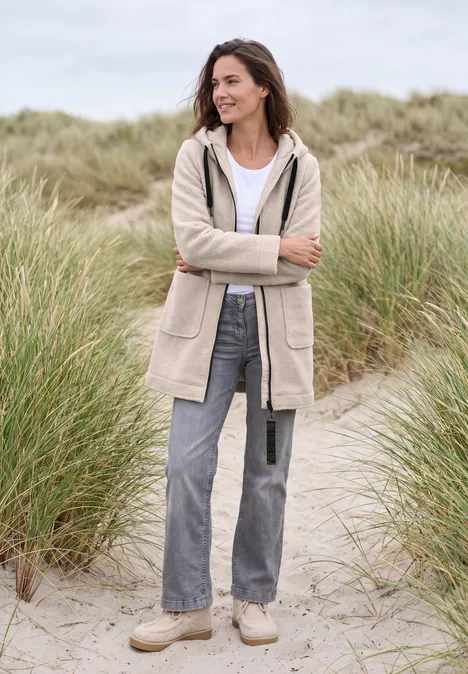 Eine lächelnde Frau steht in Stranddünen, trägt beige Jacke, Jeans und bequeme Schuhe.