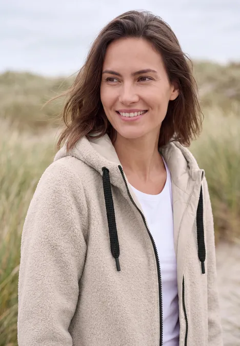 Lächelnde Frau in beiger Fleece-Jacke und weißem Shirt, im Hintergrund Dünen und Gras.