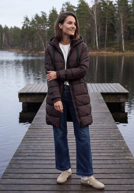 Vrouw op houten steiger bij een meer, met doorgestikte jas, jeans en sneakers, kijkt in de verte.